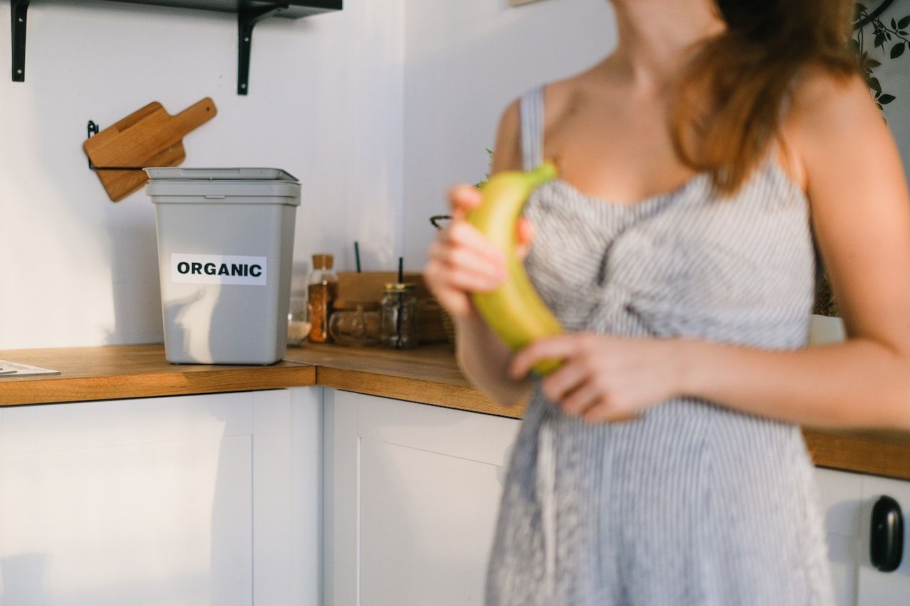 A lady holds a banana in her apartment out of focus with an organic compost bin behind her